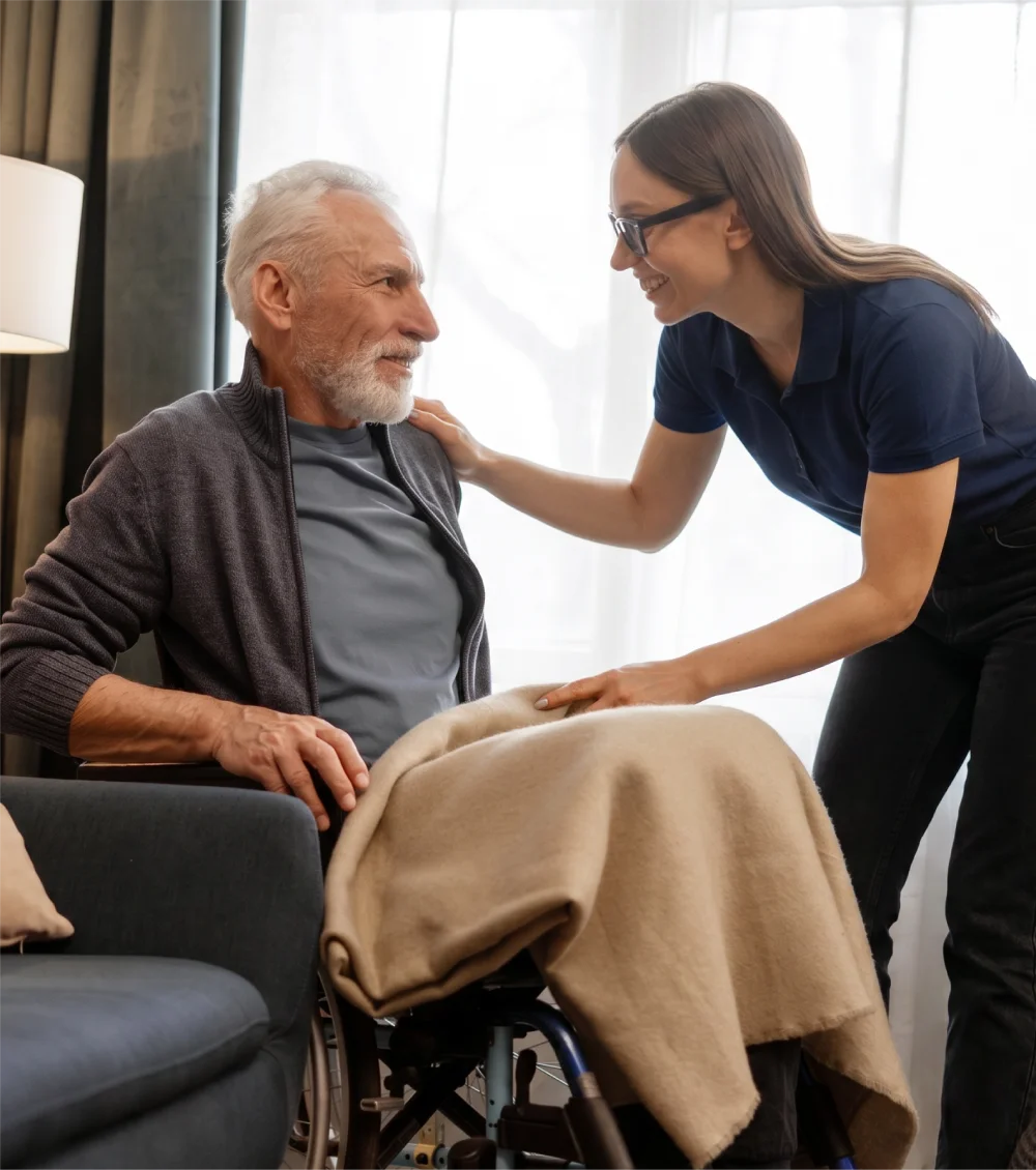 Caregiver smiling and tucking a blanket around an elderly man in a wheelchair.